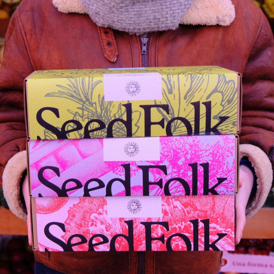 Three 'Seed Folk' boxes held by a person wearing a brown jacket.