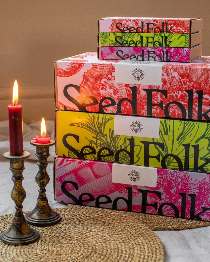 Stack of 'Seed Folk' gift boxes with candles on a textured surface